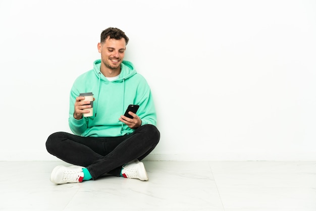 Jovem bonito homem caucasiano sentado no chão segurando um café para levar e um celular