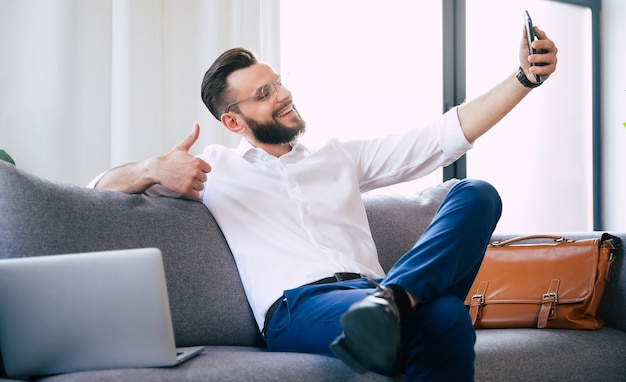 Jovem bonito homem de negócios barbudo de óculos e roupa formal elegante está sentado no sofá enquanto relaxa e usa o telefone inteligente