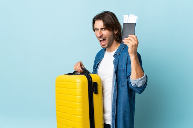 Jovem bonito isolado em um fundo azul de férias com mala e passaporte