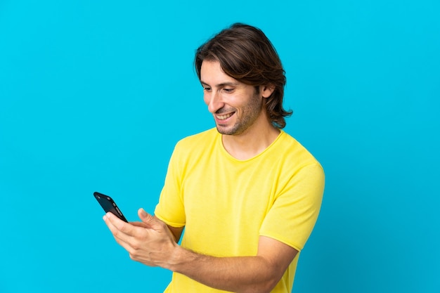 Jovem bonito isolado em um fundo azul enviando uma mensagem ou e-mail com o celular