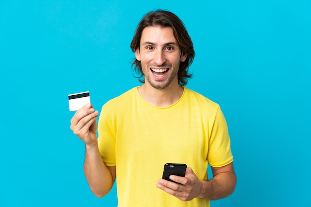 Jovem bonito isolado no azul comprando com o celular e segurando um cartão de crédito com expressão de surpresa