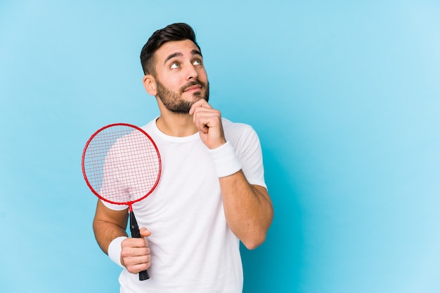 Jovem bonito jogando badminton isolado olhando de soslaio com expressão duvidosa e cética.