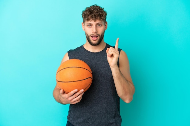 Jovem bonito jogando basquete isolado em fundo azul com a intenção de perceber a solução enquanto levanta um dedo