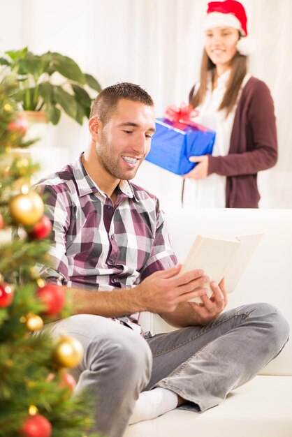 Foto jovem bonito lendo livro enquanto sua namorada segura um presente de natal e ela quer surpreendê-lo.