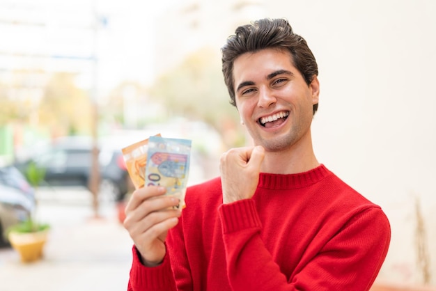 Jovem bonito levando muito dinheiro ao ar livre comemorando uma vitória