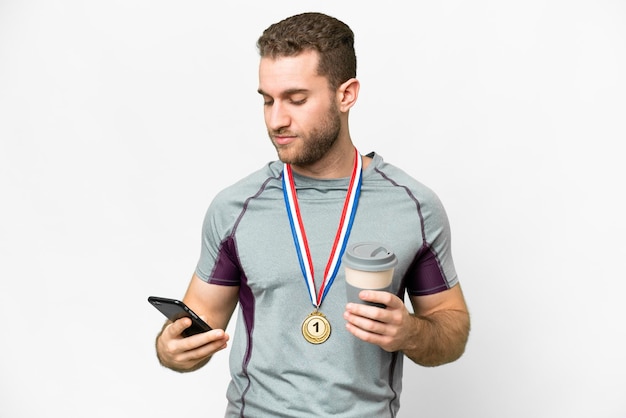 Jovem bonito loiro com medalhas sobre fundo branco isolado segurando café para levar e um celular