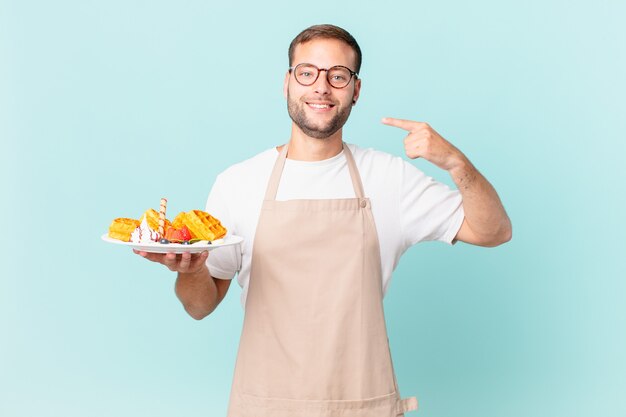 jovem bonito loiro sorrindo com confiança, apontando para o próprio sorriso largo. cozinhar waffles conceito