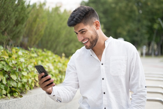 Jovem bonito mestiço sorrindo usando um telefone celular ao ar livre