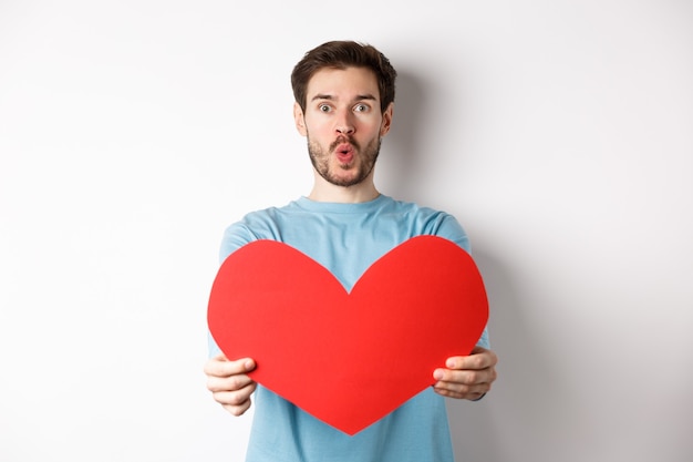 Jovem bonito mostrando um grande coração vermelho de dia dos namorados, estar apaixonado, lábios franzidos para um beijo no dia dos namorados, em pé no branco