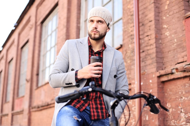 Jovem bonito no casaco cinza e chapéu e andar de bicicleta na rua bebendo café.