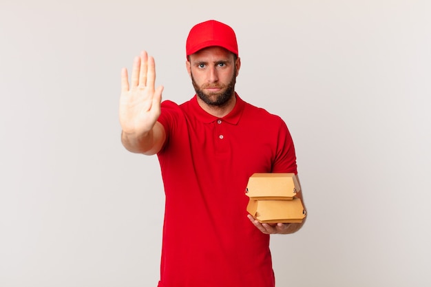 Jovem bonito olhando sério, mostrando a palma da mão aberta fazendo gesto de parada de hambúrguer entregando conceito