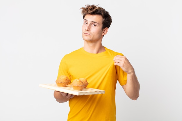 Jovem bonito parecendo arrogante, bem-sucedido, positivo e orgulhoso segurando uma bandeja de muffins