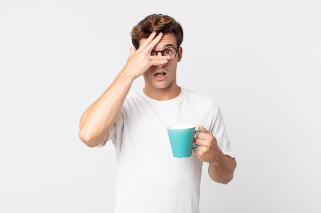 Jovem bonito parecendo chocado, assustado ou apavorado, cobrindo o rosto com a mão e segurando uma xícara de café