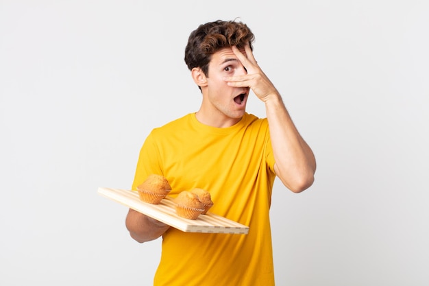 Jovem bonito parecendo chocado, assustado ou apavorado, cobrindo o rosto com a mão segurando uma bandeja de muffins