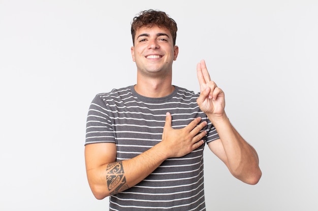 Jovem bonito parecendo feliz, confiante e confiável, sorrindo e mostrando sinal de vitória, com uma atitude positiva