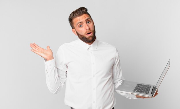 Jovem bonito parecendo surpreso e chocado, com o queixo caído segurando um objeto e segurando um laptop