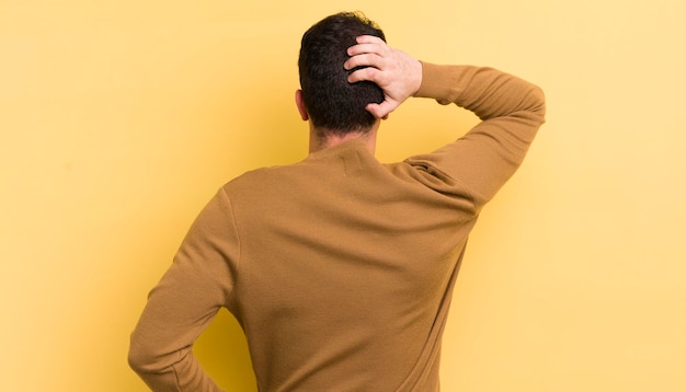 Jovem bonito pensando ou duvidando de coçar a cabeça, sentindo-se confuso e confuso na vista traseira ou traseira
