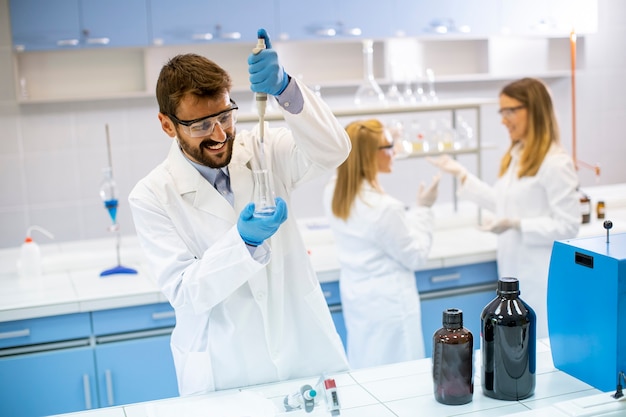 Jovem bonito pesquisador em roupas de trabalho de proteção em pé no laboratório e analisando frasco com líquido