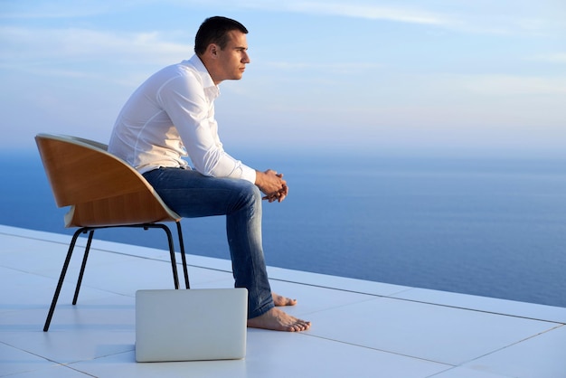 jovem bonito relaxando e trabalhando no computador portátil na varanda de casa enquanto olha o pôr do sol