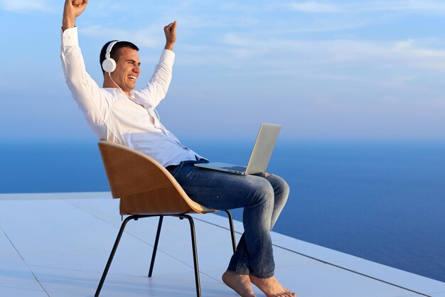 jovem bonito relaxando e trabalhando no computador portátil na varanda de casa enquanto olha o pôr do sol