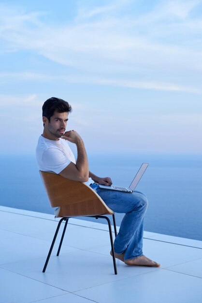 jovem bonito relaxando e trabalhando no computador portátil na varanda de casa enquanto olha o pôr do sol