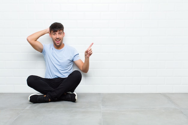 Jovem bonito rindo, olhando feliz, positivo e surpreso, percebendo uma ótima idéia apontando para o espaço lateral da cópia, sentado no chão de cimento