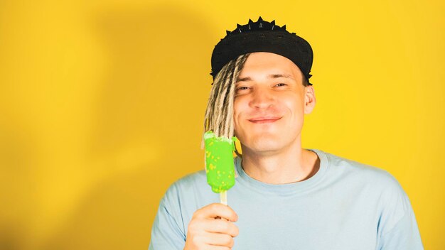 Jovem bonito satisfeito com dreadlocks no boné preto comendo sorvete no fundo amarelo Cara alegre em camiseta azul desfrutando de sundae