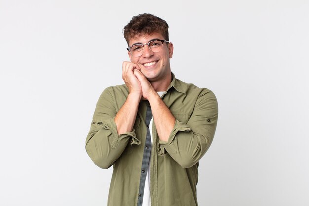 Jovem bonito se apaixonando e parecendo fofo, adorável e feliz, sorrindo romanticamente com as mãos ao lado do rosto