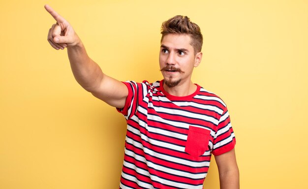 Jovem bonito se sentindo chocado e surpreso apontando e olhando para cima com admiração com o conceito de bigode estranho de boca aberta espantado