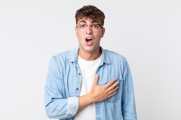 Jovem bonito se sentindo chocado e surpreso, sorrindo, levando a mão ao coração, feliz por ser o único ou mostrando gratidão