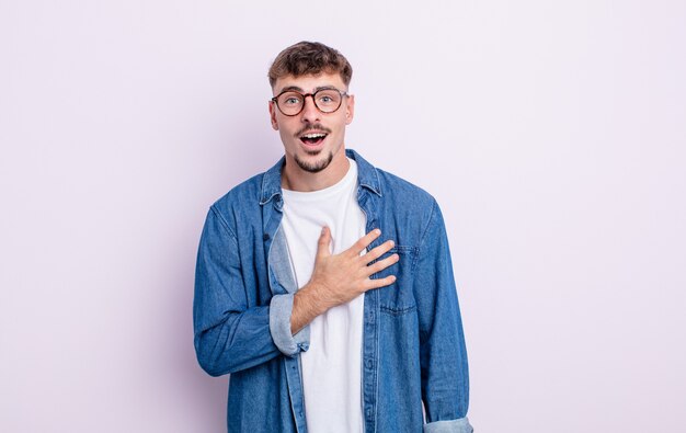 Jovem bonito se sentindo chocado e surpreso, sorrindo, levando a mão ao coração, feliz por ser o único ou mostrando gratidão