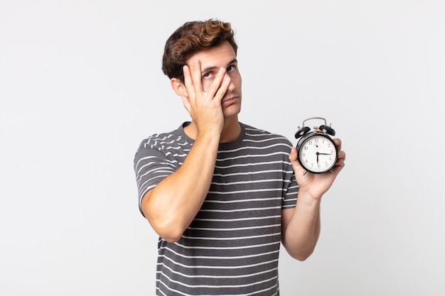 Jovem bonito se sentindo entediado, frustrado e com sono depois de um dia cansativo segurando um despertador