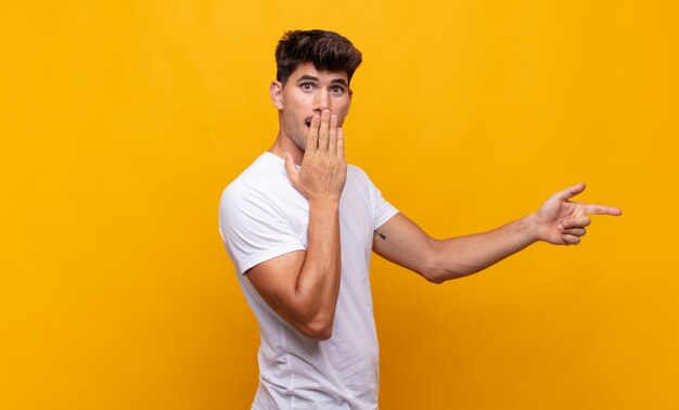 Jovem bonito se sentindo feliz, chocado e surpreso, cobrindo a boca com a mão e apontando para o espaço lateral da cópia