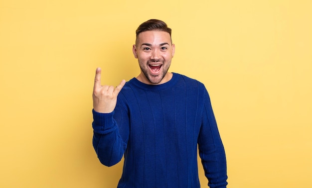 Jovem bonito se sentindo feliz, divertido, confiante, positivo e rebelde, fazendo sinal de rock ou heavy metal com a mão