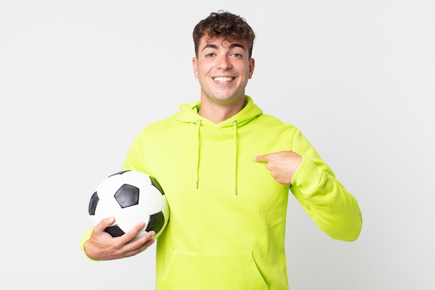 Jovem bonito se sentindo feliz e apontando para si mesmo com um animado e segurando uma bola de futebol