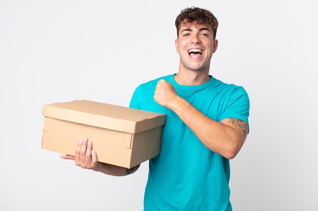 Jovem bonito se sentindo feliz e enfrentando um desafio ou comemorando e segurando uma caixa de papelão