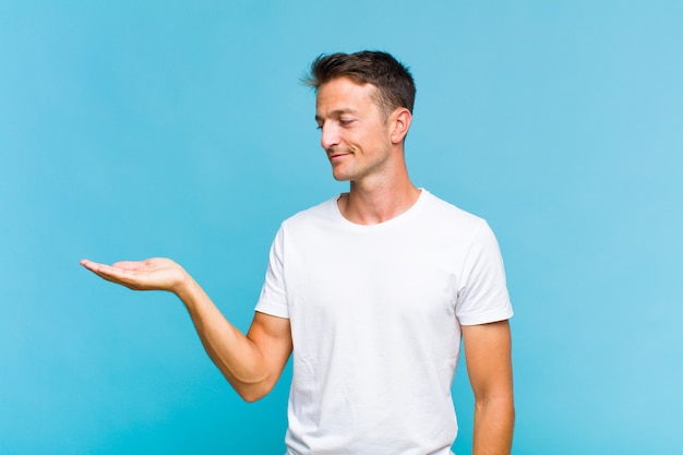 Jovem bonito se sentindo feliz e sorrindo casualmente, olhando para um objeto ou conceito seguro ao lado