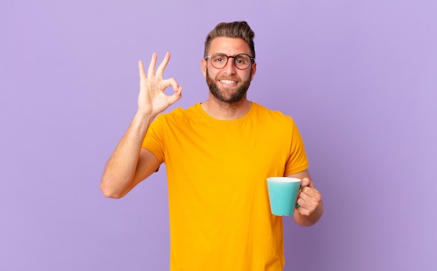 Jovem bonito se sentindo feliz, mostrando aprovação com um gesto certo. e segurando uma caneca de café