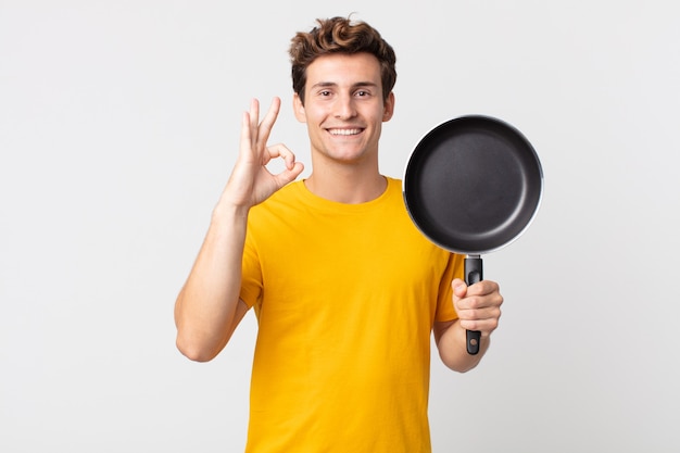 Jovem bonito se sentindo feliz, mostrando aprovação com um gesto de ok e segurando uma panela