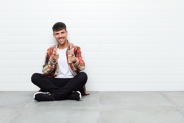 Jovem bonito se sentindo feliz, tranquilo, satisfeito, relaxado e bem-sucedido, apontando, escolhendo você sentado no chão de cimento