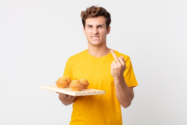 Jovem bonito se sentindo irritado, irritado, rebelde e agressivo segurando uma bandeja de muffins