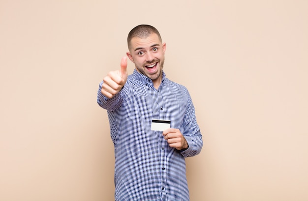 Jovem bonito se sentindo orgulhoso, despreocupado, confiante e feliz, sorrindo positivamente com o polegar para cima com um cartão de crédito