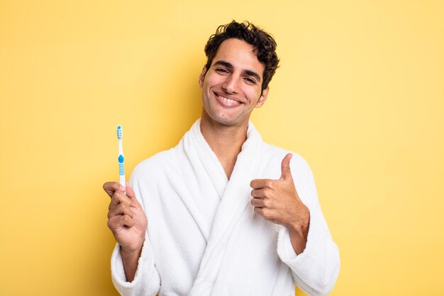 Jovem bonito se sentindo orgulhoso, sorrindo positivamente com o polegar para cima. conceito de roupão e escova de dentes