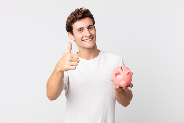 Jovem bonito se sentindo orgulhoso, sorrindo positivamente com o polegar para cima e segurando um cofrinho