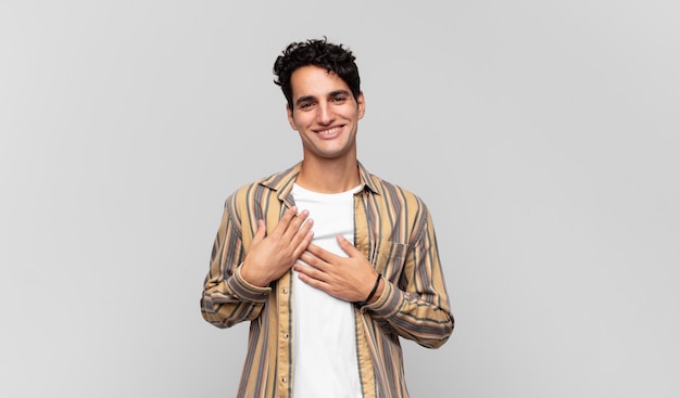 Jovem bonito se sentindo romântico, feliz e apaixonado, sorrindo alegremente e segurando as mãos perto do coração