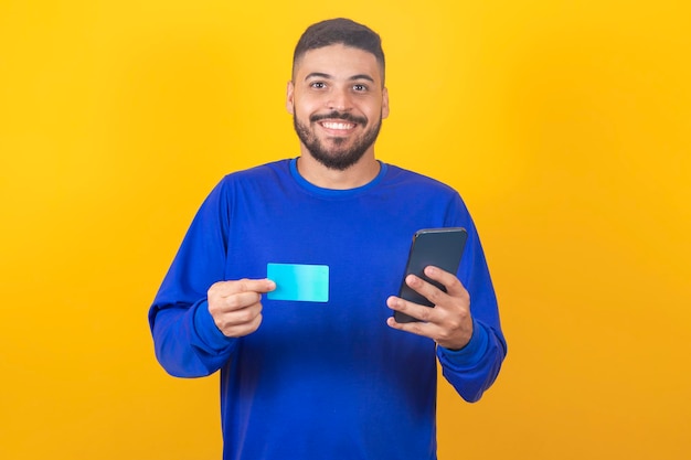 Jovem bonito segura cartão de crédito e smartphone isolado em fundo amarelo Compras online Pagamento online