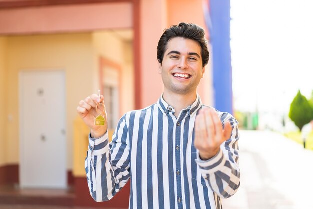 Jovem bonito segurando as chaves de casa ao ar livre convidando para vir com a mão Feliz por você ter vindo