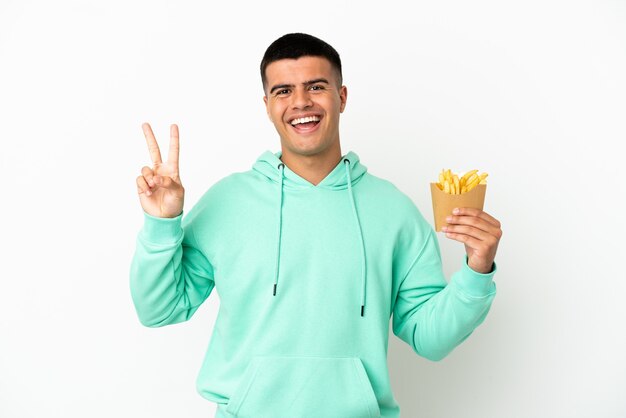 Jovem bonito segurando batatas fritas sobre uma parede branca isolada, sorrindo e mostrando o sinal da vitória