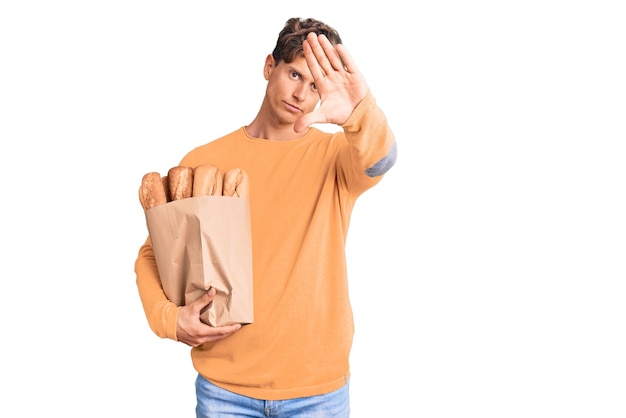 Jovem bonito segurando o saco de papel com pão com a mão aberta, fazendo o sinal de pare com gesto de defesa de expressão séria e confiante