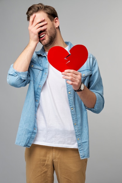 Foto jovem bonito segurando papel quebrado coração vermelho dos namorados em pé sobre o cinza